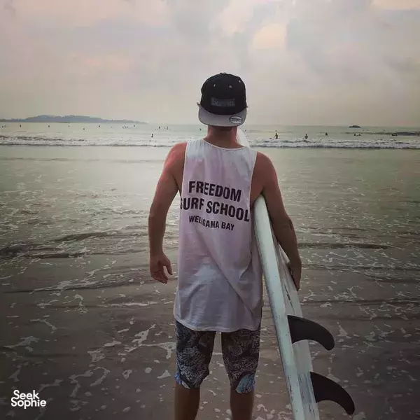 Trainee in a Freedom Surf School Weligama Bay tank top standing at the beach with a surfboard, facing the waves
