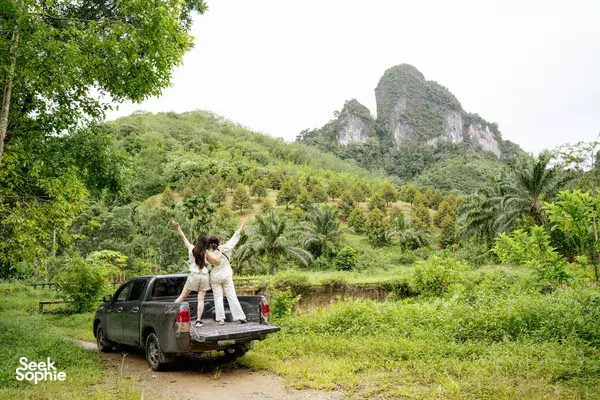 Rider standing in the back of a pickup truck on a dirt road through jungle greenery and limestone hills in Thailand