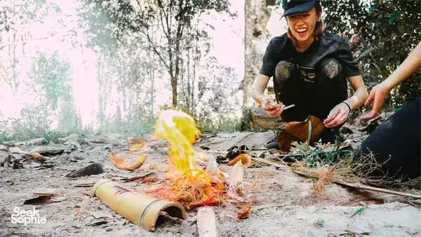 Smiling jungle survival participant stirring food over an open fire in a forest clearing in Malaysia, wearing a dark T-shirt