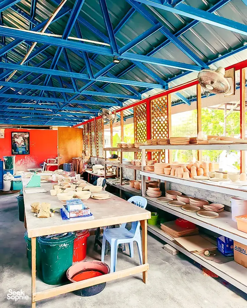 Wide view of Ilham Ceramic Studio in Langkawi, with blue rafters, rows of ceramics on racks, and tools on benches