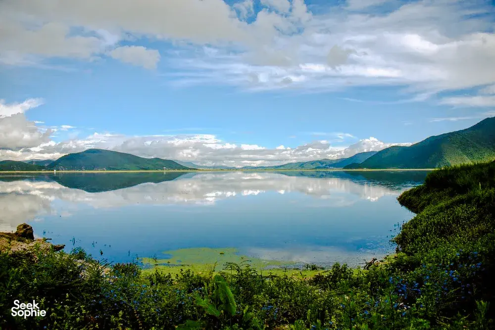 Calm Napa Lake in Shangri-La framed by forested hills and mountains under cloudy skies, with mirror-like reflections