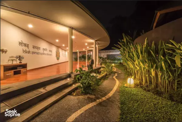Warm-lit yoga studio corridor at night with a pathway beside plants and lit practice space outside, in Indonesia
