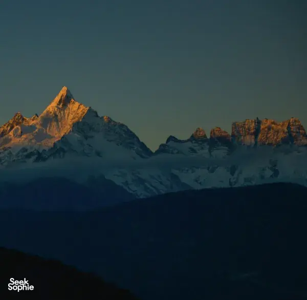 Sunlit snow-capped mountain peaks above dark ridgelines in Shangri-La at dusk, with a dramatic clear sky fading to blue