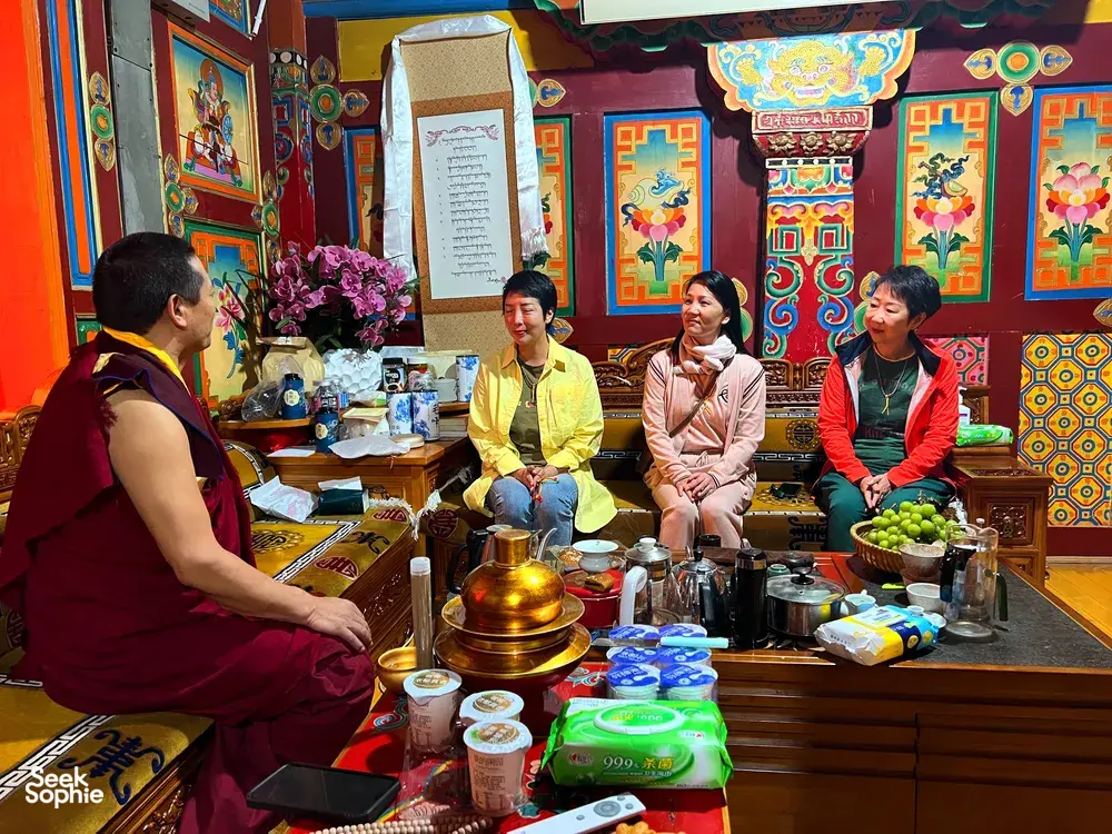 Lama in maroon robes leads a tea-time conversation as travellers sit by a decorated table of pots in Shangri-La