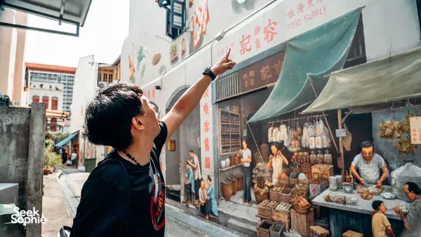 Triad Trails guide pointing along a narrow Singapore market lane lined with curtained stalls and food-shop displays