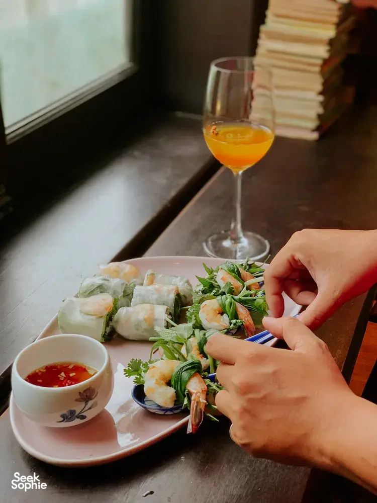 Tangan sedang meracik lumpia segar dengan herba dan saus cocolan di Chan Dat Living Room, Hoi An, ditemani minuman berwarna oranye
