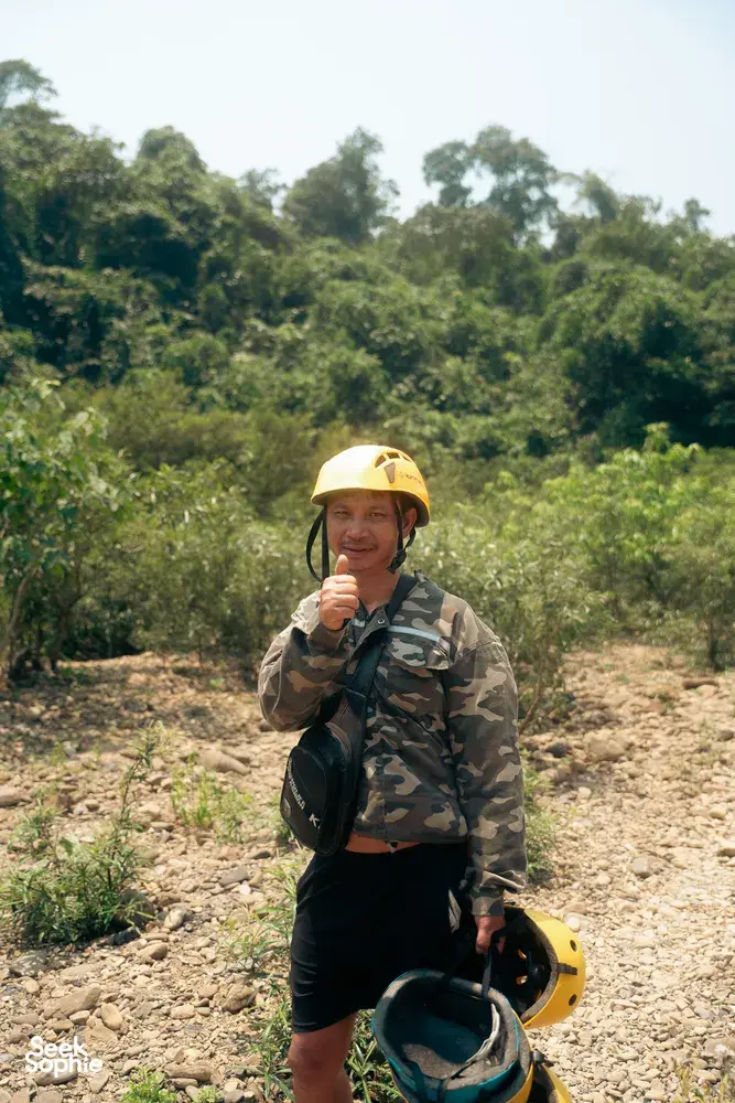 Traveller in a yellow helmet and camouflage stands on a rocky trail through Phong Nha jungle, holding up a snack