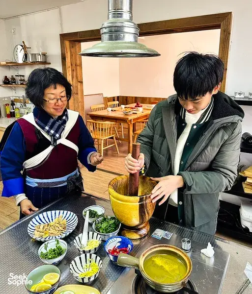 In a home kitchen in Lijiang, a host guides a guest while mixing ingredients in a jar during Yunnan cooking
