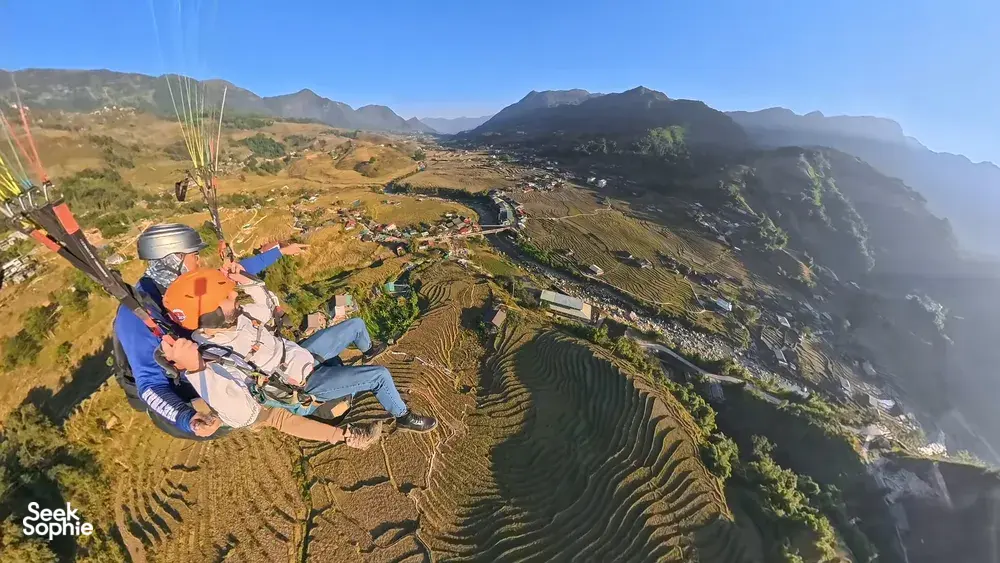 Orange-wing paraglider seated in harness, drifting over zigzag terraced rice plots and mountain slopes with Sapa Paragliding above Sapa