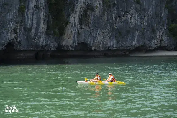 Kayakers aboard a Cat Ba boutique cruise paddle across calm green waters in Lan Ha Bay beside limestone karst cliffs
