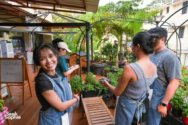 Two guests in aprons chat on a garden patio with plants and seating, beginning private pizza making at Secret Pizza Club in Singapore