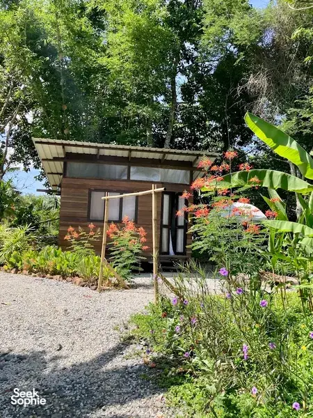 Wood-and-glass cabin at Kebun Lidstari farmstay in Langkawi, tucked into a colourful tropical garden with blooms and banana leaves