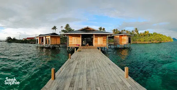Wooden stilt sea lodges on a tropical island coastline, with a walkway leading out over clear water in Semporna