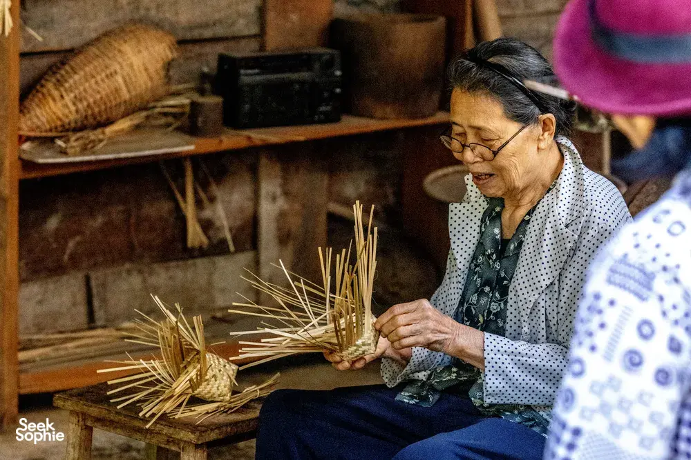 Woman sits in a rustic kitchen workshop, braiding dried straw or pine needles for ingredients in a Mae Kampong forage-to-table class