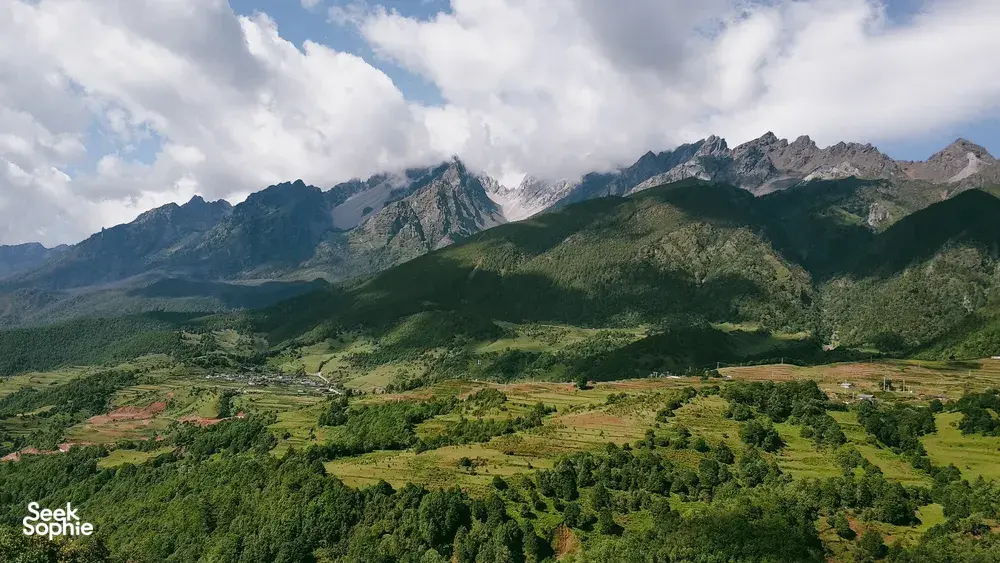Lush green landscape around steep peaks near Lijiang, viewed from above with scattered clouds drifting over the mountains