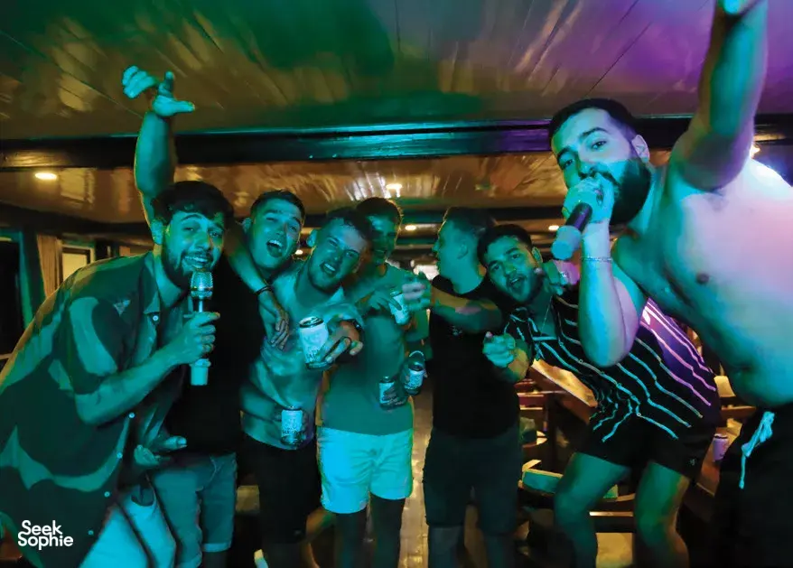 Group of friends cheering and drinking on board Oasis Bay Party Cruise at night under neon lighting, with drinks in hand in Halong Bay