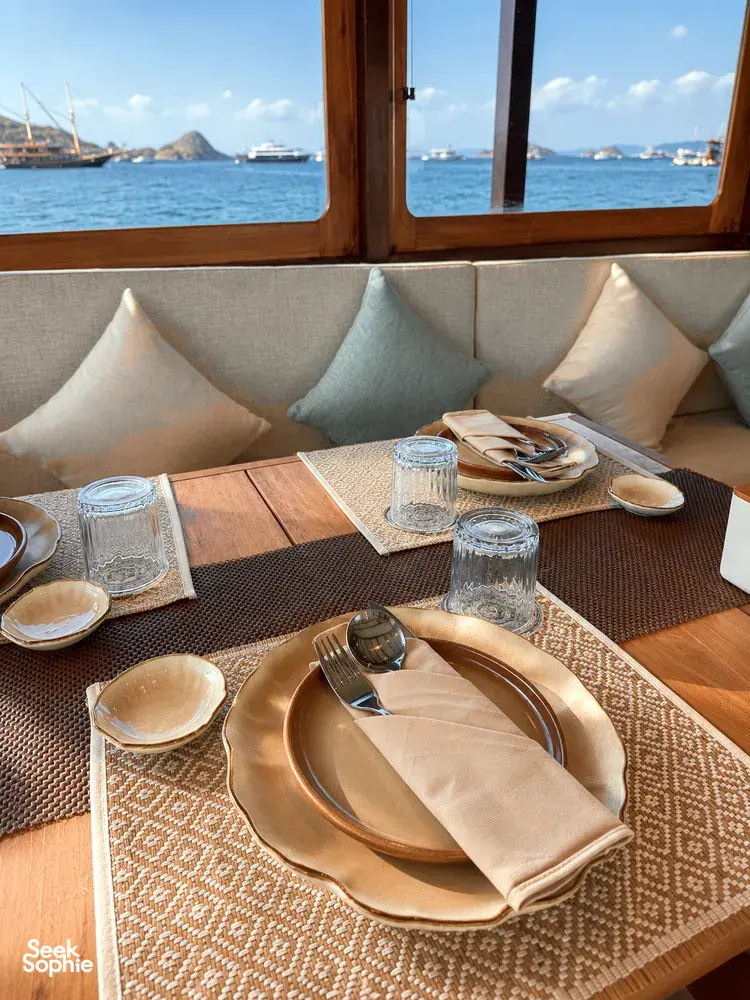 Breakfast table set on a phinisi in Labuan Bajo, with plates, cutlery and patterned placemats by large sea windows