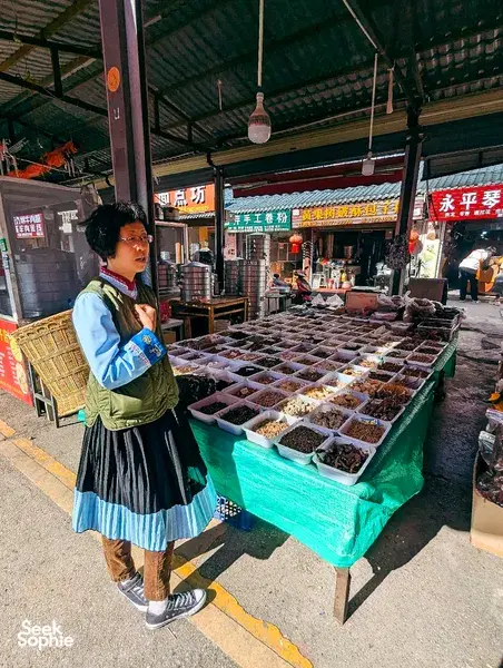 Fancy in traditional Naxi dress at a Lijiang market stall, standing by trays of dried herbs and spices on a morning walk