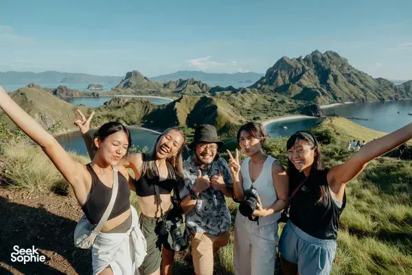 Group of travellers laughing with hands raised on a mountain ridge above a winding bay, rugged peaks and water
