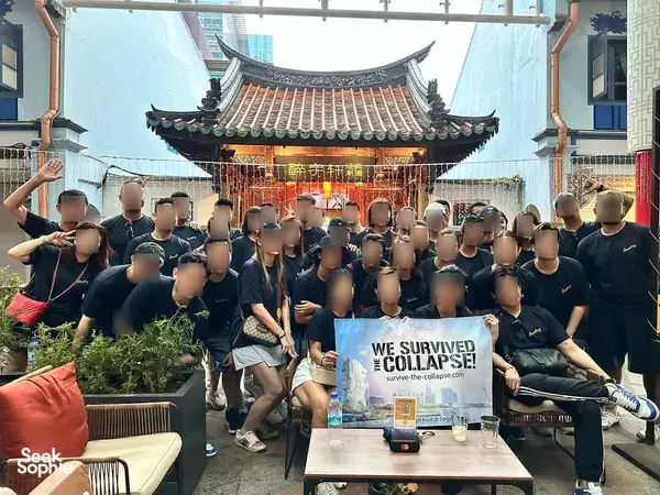 Blurred-head group poses in a temple courtyard beside a banner reading 'We Survived the Collapse!' for team building Singapore