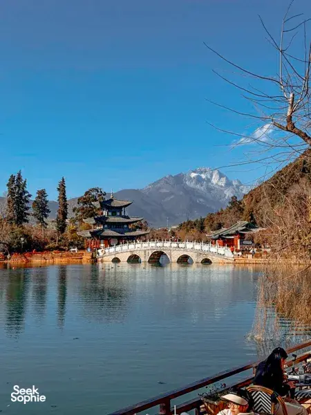 Red arched bridge and traditional pavilion on a calm lake, with snow-capped mountains behind it in Lijiang