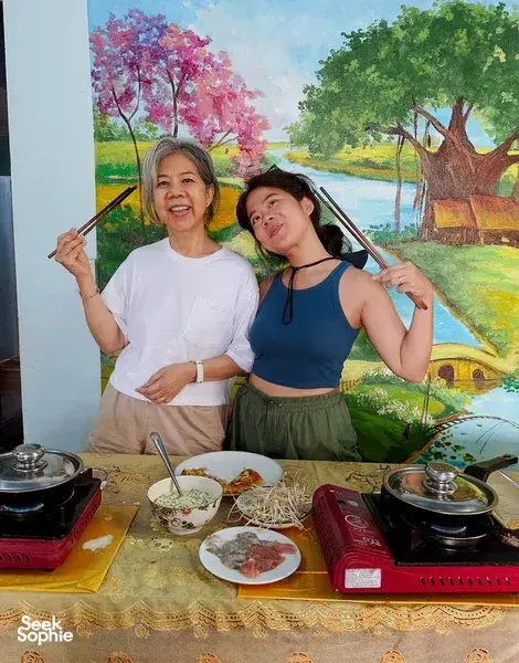 Hoi An countryside home-cooked meal: two travellers pose beside bowls of noodles and hotpots on a covered family table