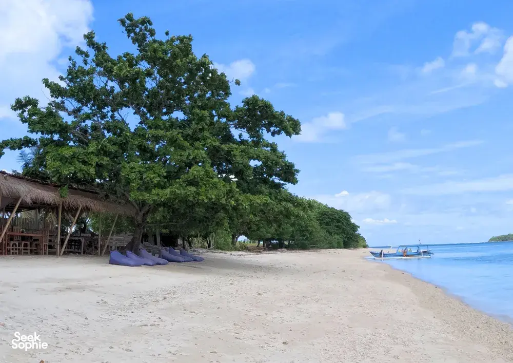 Pantai yang tenang di Kepulauan Gili, Lombok, dengan pondok beratap rumbia, air yang tenang, dan sebuah bot kecil yang sedia untuk sesi snorkel peribadi