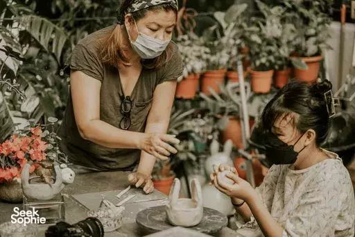 Masked instructor helping a child water and pot flowers at a bench surrounded by potted plants in Singapore during workshops in Singapore