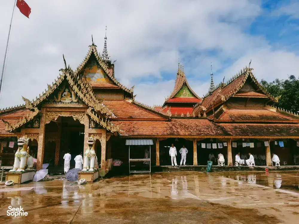 Bangunan kuil yang megah di Xishuangbanna dengan atap berlapis emas, patung, dan halaman yang basah oleh hujan di bawah langit mendung