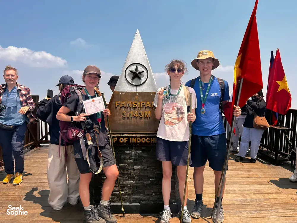 Trek group posing beside the Fansipan summit marker at 3,143m with flags on a platform on Mt Fansipan in Sapa
