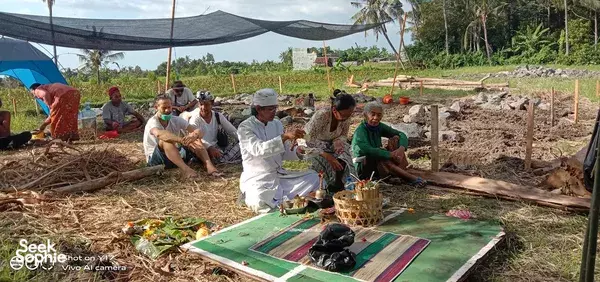 Priest and guests seated on woven rugs with ceremonial offerings outdoors at Omunity Bali in Bali during a cleansing ceremony
