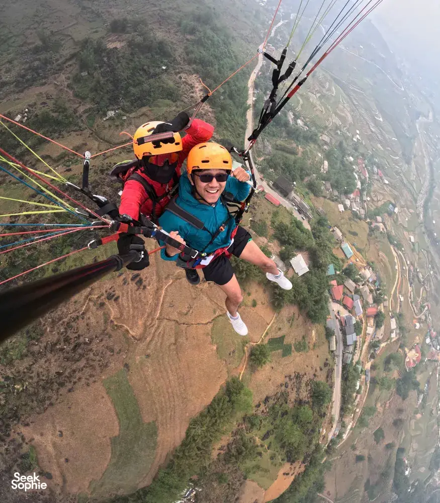 Smiling paraglider passenger in a helmet glides high above green hills and river valleys with gear visible in Sapa