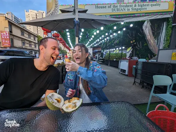 Travellers tasting durian at a busy Geylang night market stall in Singapore, smiling with wedges under hanging lights