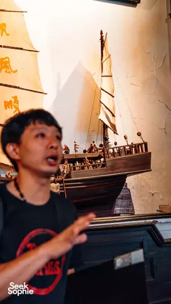 Triad Trails guide gesturing beside a wooden model ship display inside a Singapore heritage room on a gritty past walk