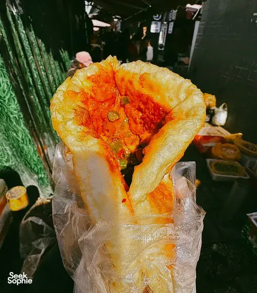 Stuffed golden flatbread sprinkled with red chilli spices held over wrapping at a lively Lijiang night market snack stall