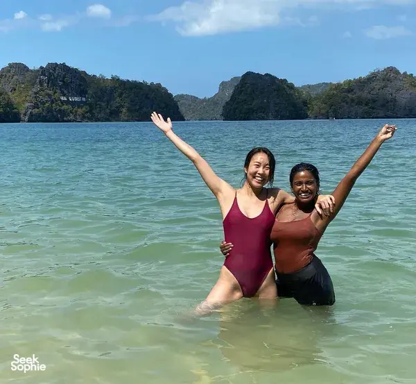 Travellers cheer with arms raised in shallow sea water off Langkawi, with green island cliffs rising in the background