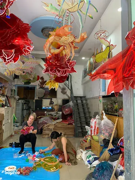 Traditional lantern workshop in Saigon with red and gold paper lanterns hanging overhead as makers work at tables in the shop