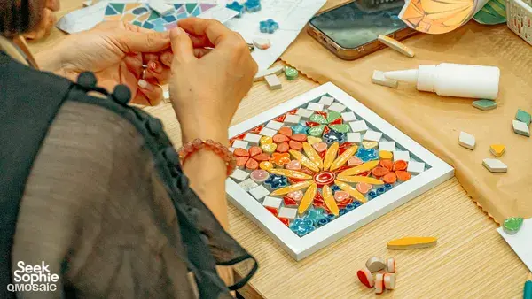 Hands placing small ceramic pieces into a bright star mosaic on a board during a QMosaic Artspace & Workshop session in Bat Trang near Hanoi