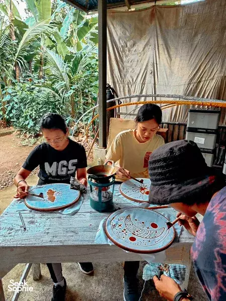 People painting decorative plates at a community workshop in a tropical setting in Indonesia, seated at tables amid greenery