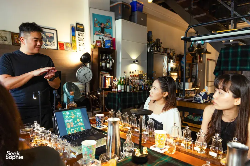Small-batch whisky tasting in a hidden home bar in Singapore, a host pours for guests at a table of sample glasses and bottles