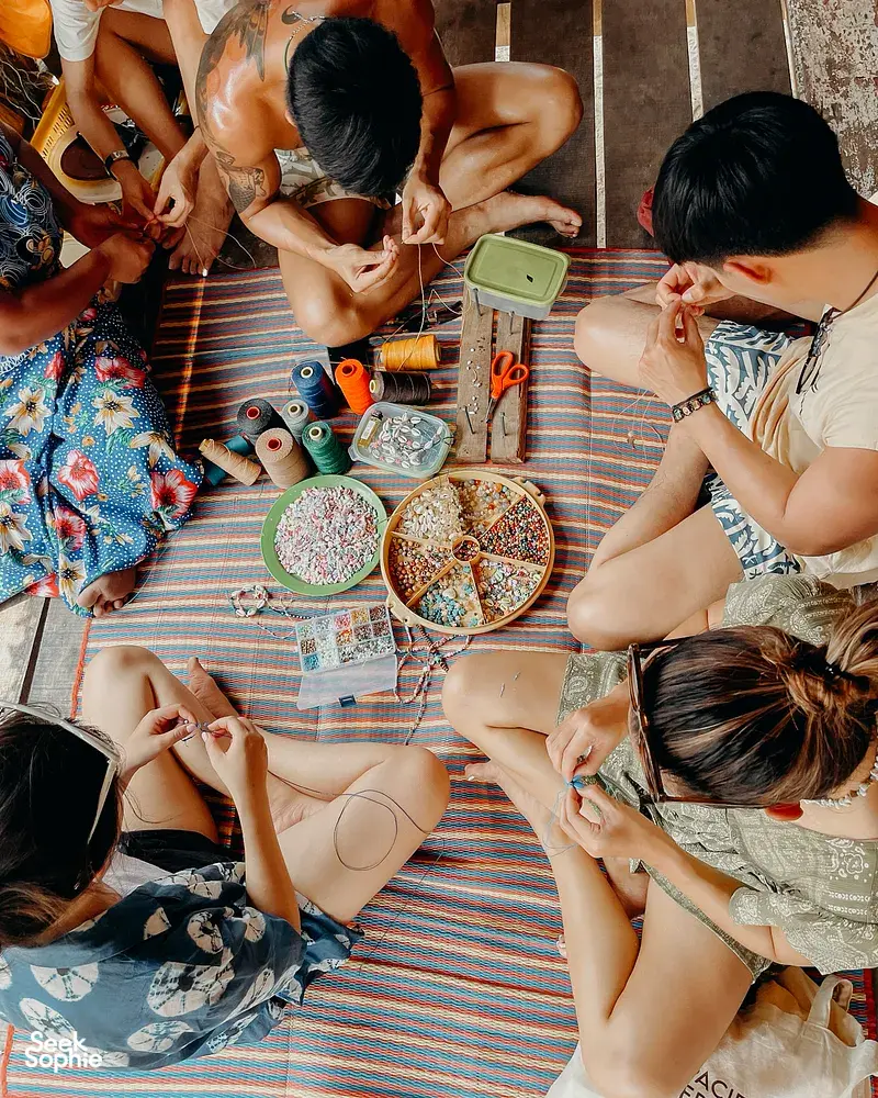 Participants making bead bracelets while seated on a woven mat inside a wooden hut during the 2D1N Surin Island trip