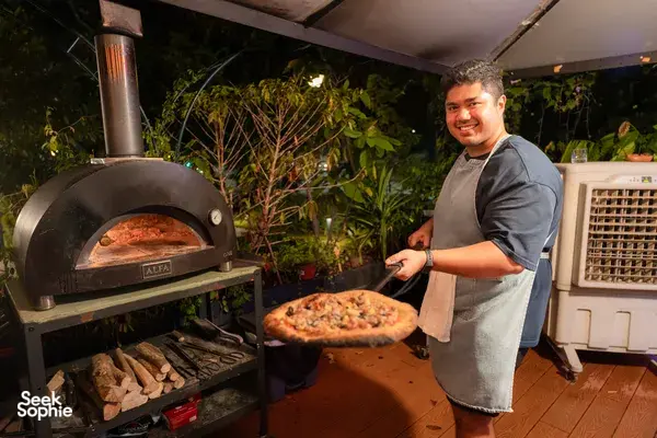 Host tending the wood-fired oven at Secret Pizza Club rooftop in Singapore, smiling beside a paddle with a fresh pizza