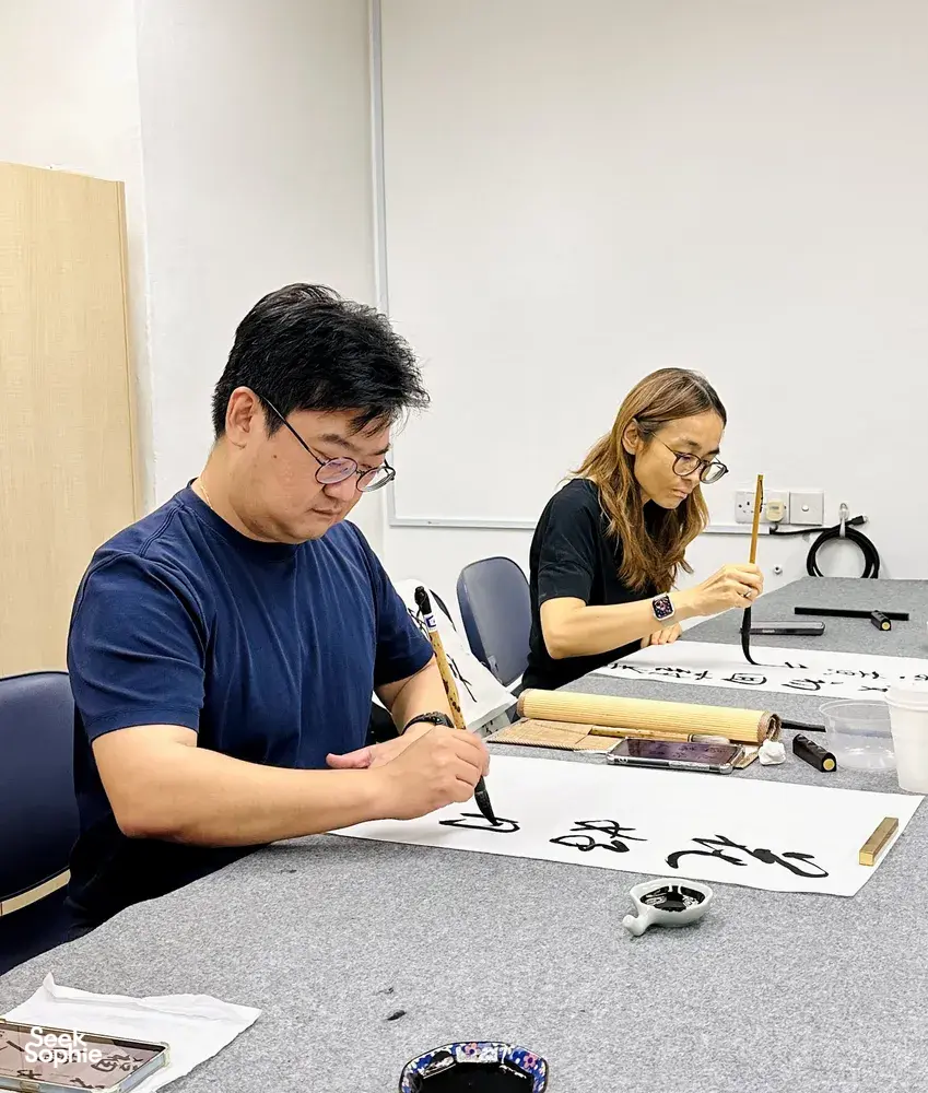 Chinese calligraphy workshop in Singapore with two learners writing on rice paper at tables, brushes and ink ready