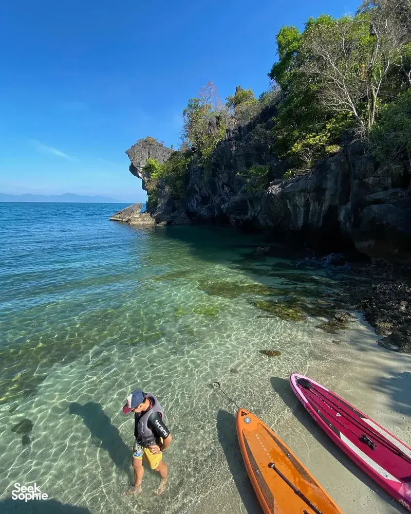 A paddler wades beside a maroon SUP board in shallow clear water along a forested Langkawi shoreline with rocky headland