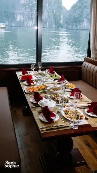 Set meja makan dengan hidangan makanan laut di kabin pelayaran Cat Ba, dibingkai tingkap tinggi yang menghadap teluk berkabus