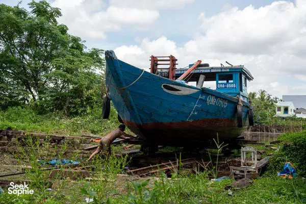 Blue wooden boat pulled up on land among weeds and greenery in Hoi An, ready for the river journey ahead