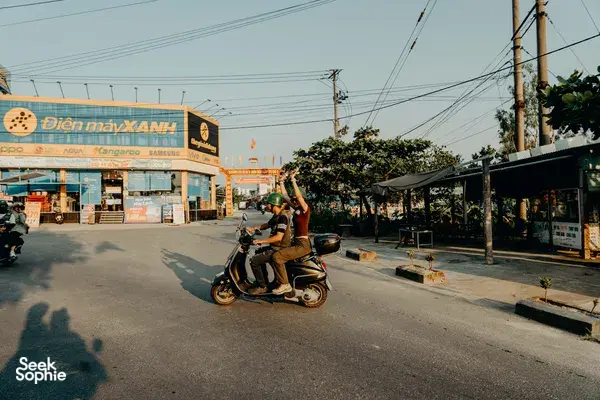 Two riders on a Vespa raise their hands as they drive past shops in Hoi An during a fun guided backroad trip