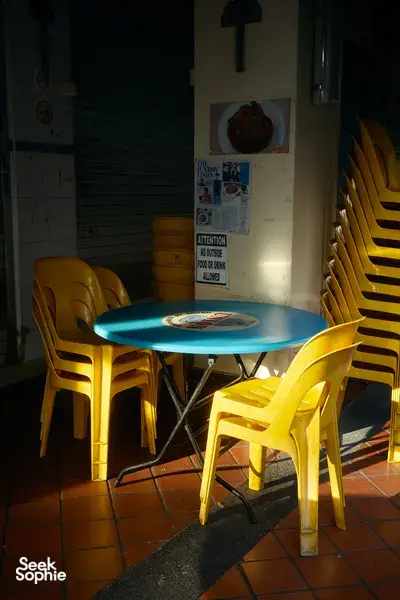 Sunlit café patio table and yellow chairs beside notices on a wall in Eastside Joo Chiat, Singapore, courtyard