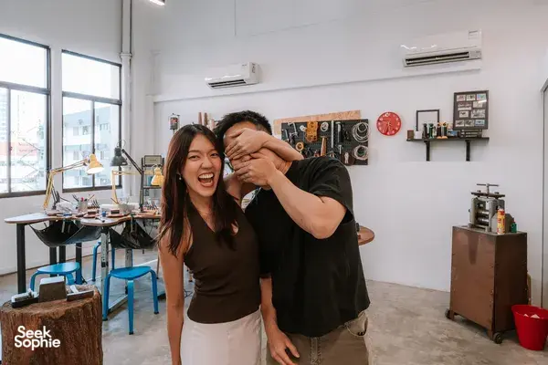 Laughing couple posing in a bright silversmithing studio with workbenches and tools behind them at Fat Anvil Studios, Singapore