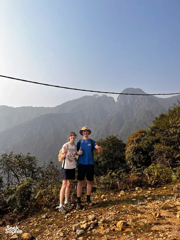 Two hikers pause on a mountain trail above Sapa as ridgelines rise behind them, towards Mt Fansipan